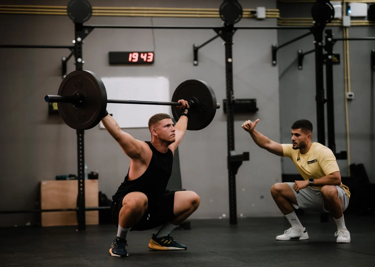 A muscular man performing a weightlifting squat with a coach guiding him indoors.